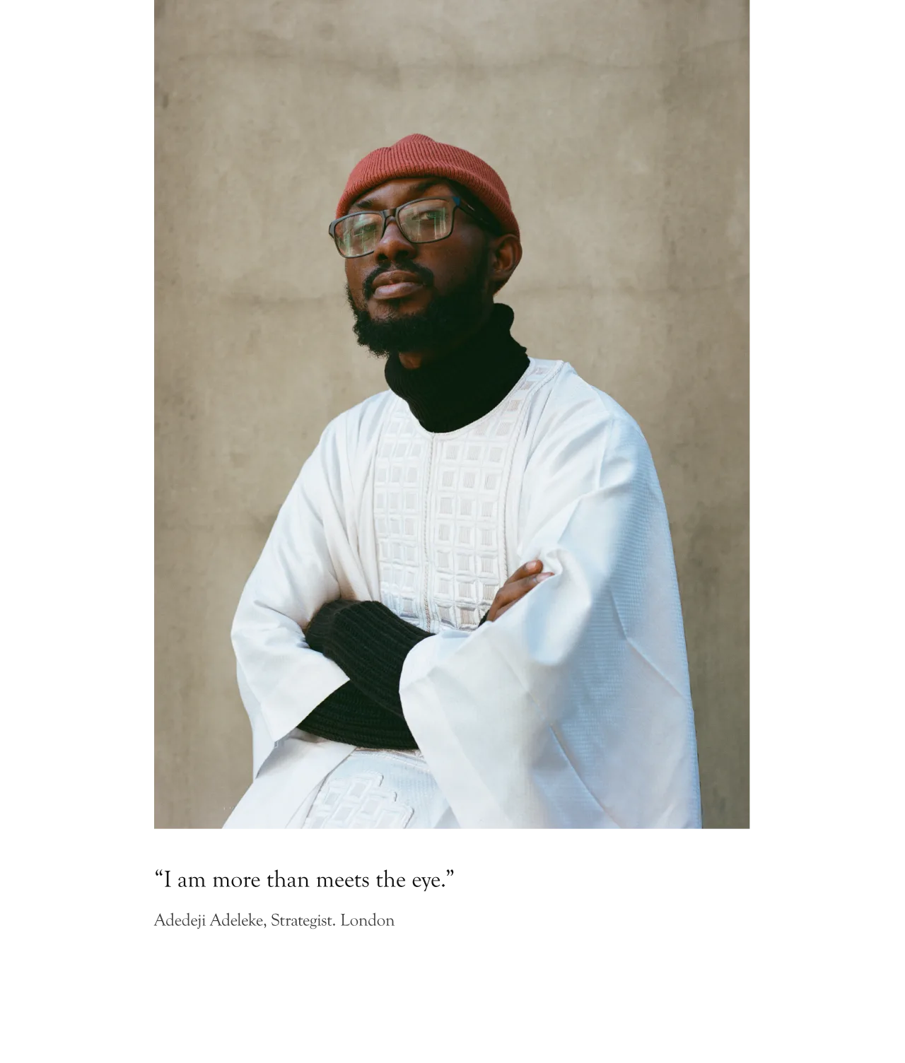 A man wearing glasses, a red knit hat, a black turtleneck, and a white patterned traditional robe sits with arms crossed, looking confidently at the camera against a plain beige background.