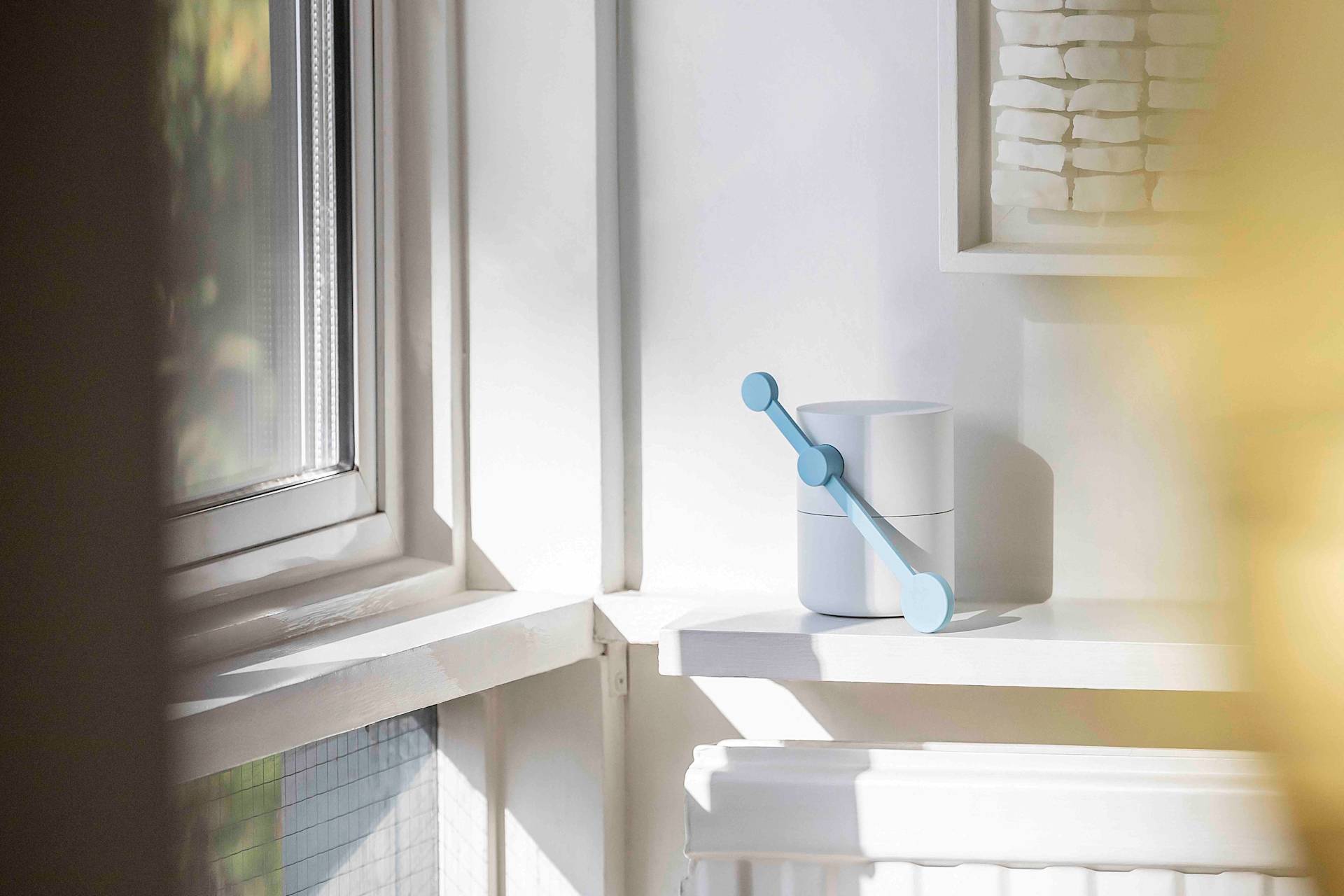 A modern white container with a blue handle sits on a sunlit windowsill, next to a white wall and radiator, with part of a framed artwork visible in the background.