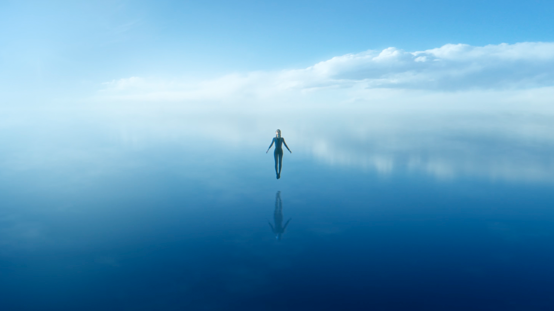 A person stands calmly in the center of a vast, still body of water, surrounded by a clear blue sky and soft clouds reflected on the waters surface, creating a serene and tranquil scene.