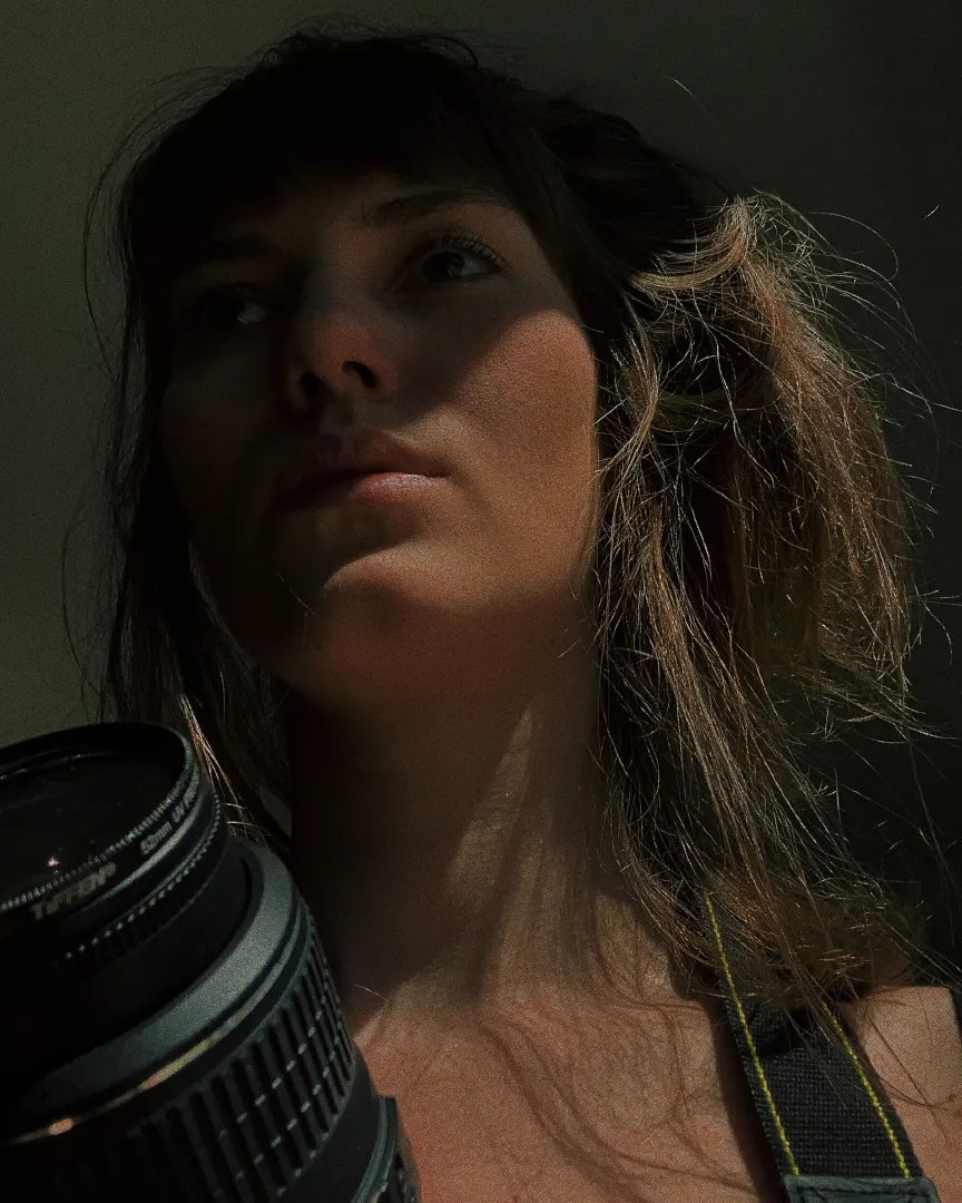 A woman with tousled hair looks thoughtfully into the distance, partially lit by natural light and shadows. She holds a camera, and wears a camera strap over her shoulder.