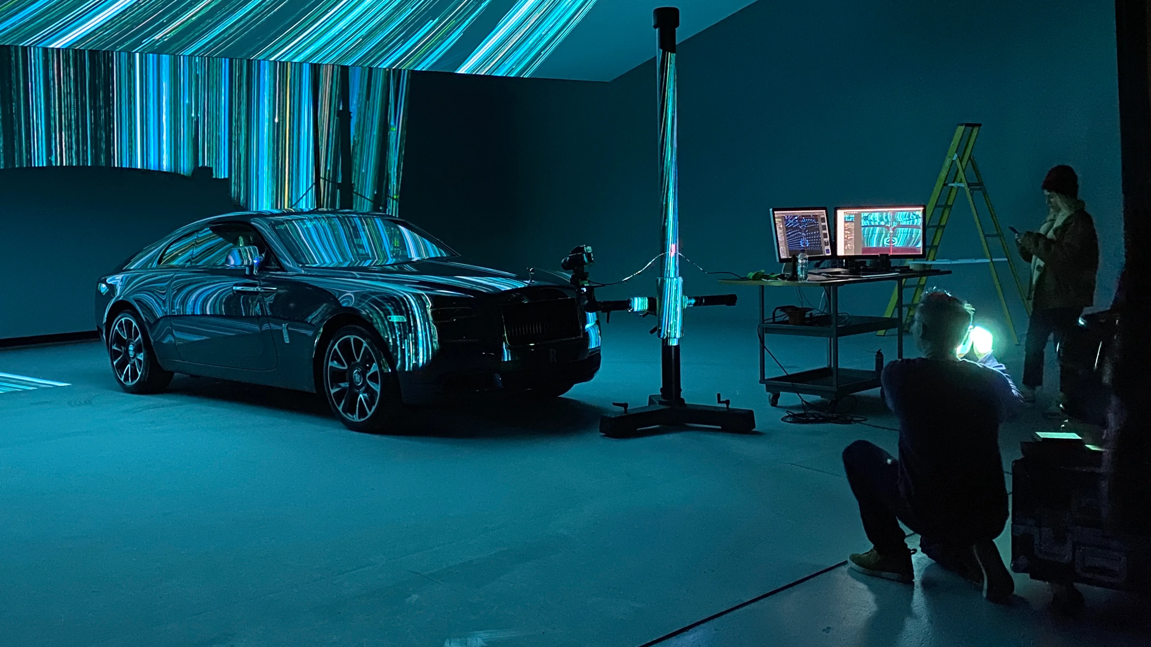 A photographer kneels to take photos of a black car in a studio with blue and green light projections. Two people stand near monitors and a ladder, surrounded by equipment. The scene has a high-tech, creative atmosphere.
