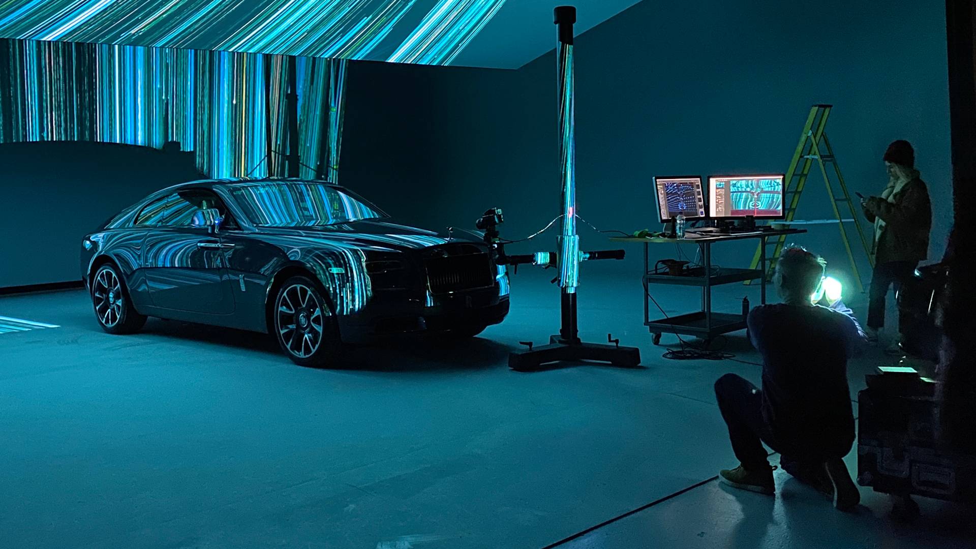 A photographer kneels to take photos of a black car in a studio with blue and green light projections. Two people stand near monitors and a ladder, surrounded by equipment. The scene has a high-tech, creative atmosphere.