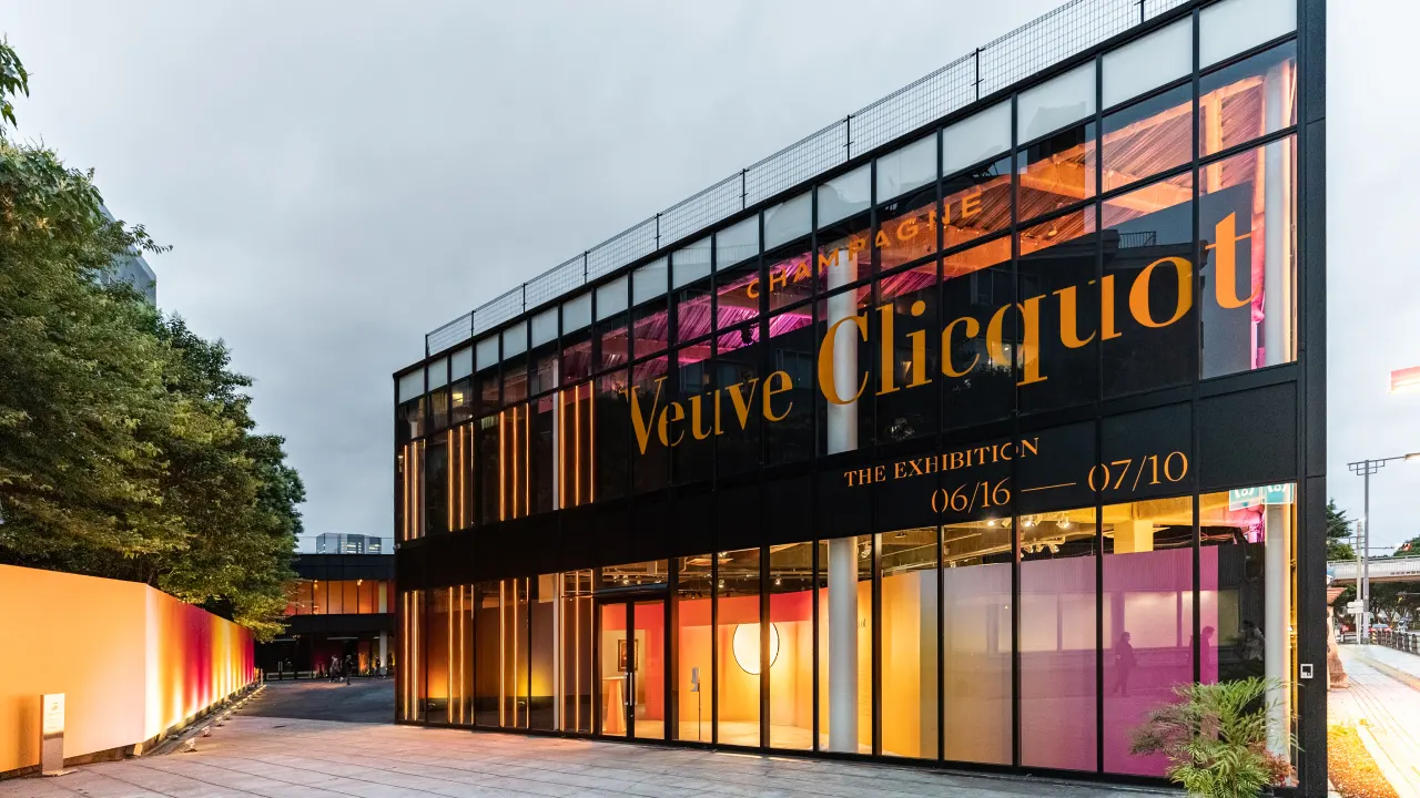 A modern glass building with “Veuve Clicquot” and exhibit dates (06/16—07/10) in large yellow letters on the facade; colorful lighting and trees are visible around the entrance.