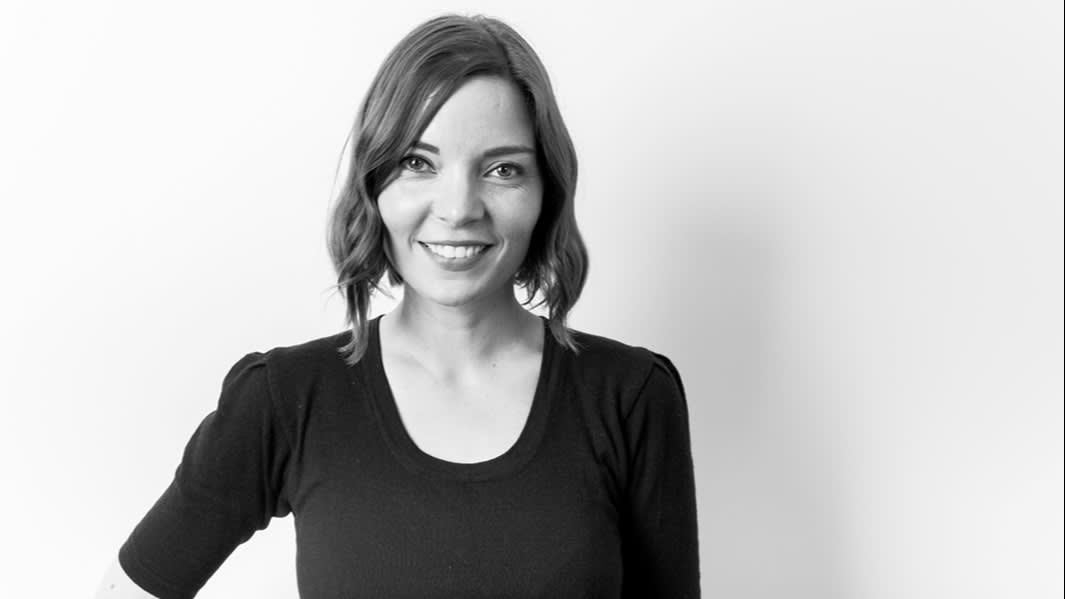 A woman with shoulder-length hair smiles at the camera. She is wearing a dark, fitted top with three-quarter sleeves and stands against a plain, light-colored background in this black and white photo.