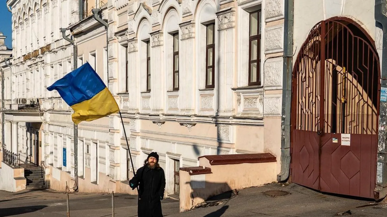 A person in dark clothing stands on a street holding a large Ukrainian flag, with ornate white buildings and a tall metal gate in the background, in bright daylight.