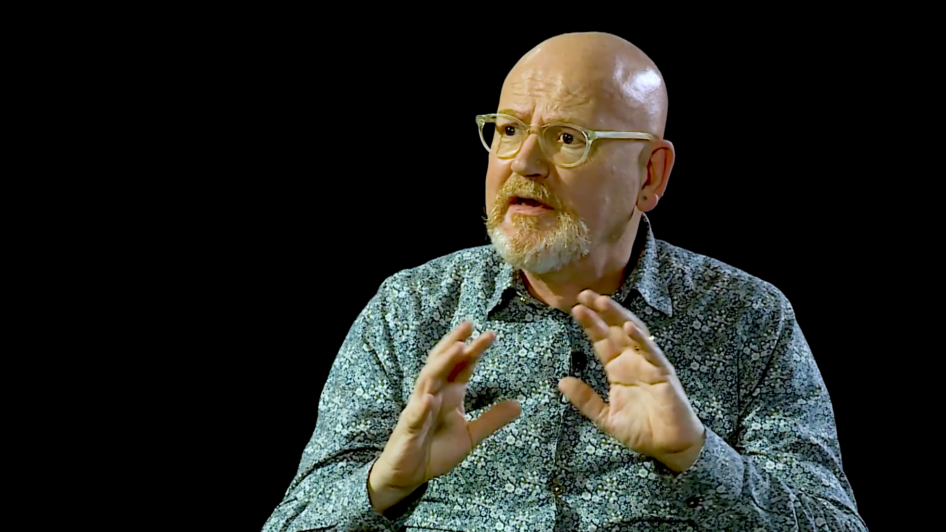 A bald man with glasses and a beard, wearing a patterned shirt, gestures with his hands while speaking against a black background.