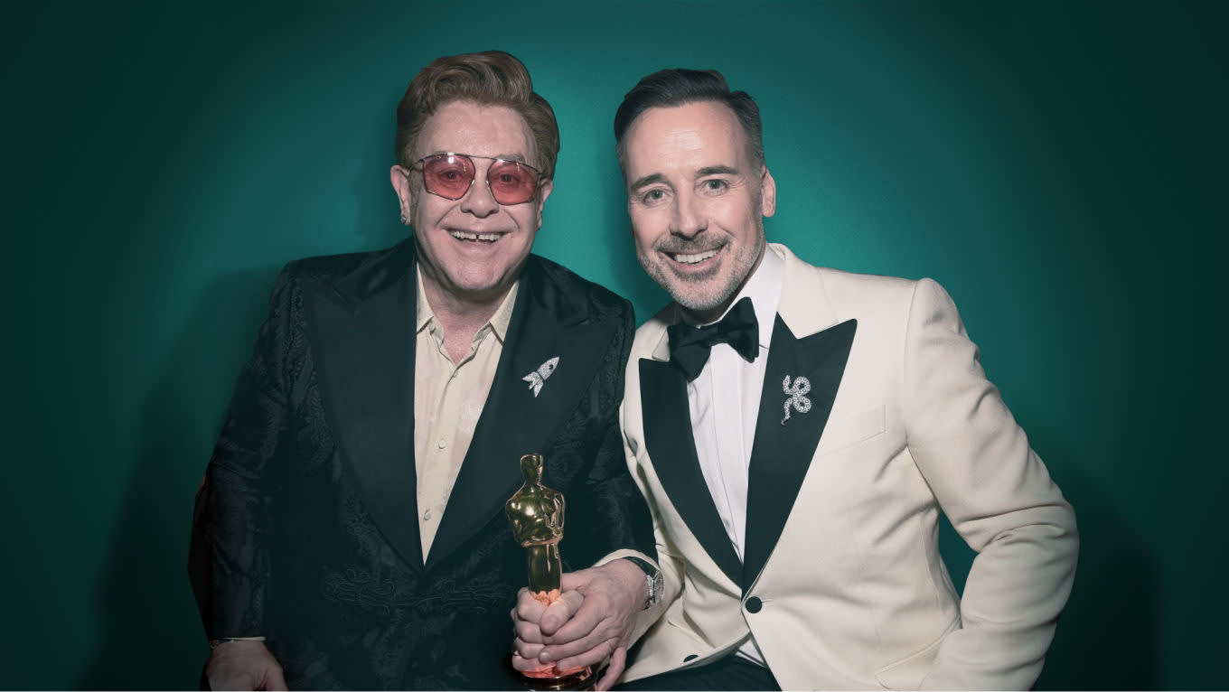 Two men pose together, smiling in front of a teal background. One wears sunglasses and a dark suit, holding a gold Oscar statuette. The other wears a white tuxedo jacket with a black lapel and bow tie.