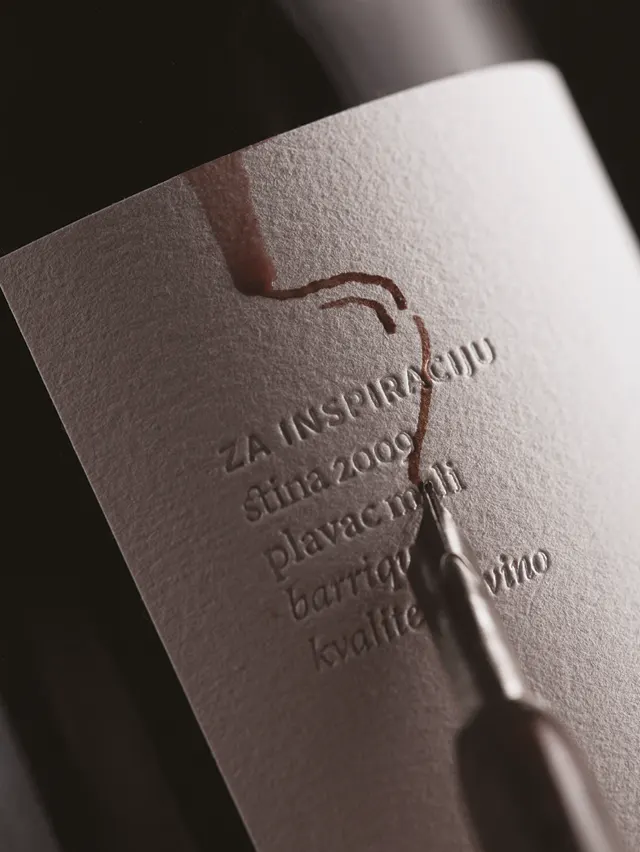 A close-up of a wine bottle label with embossed text, featuring a spill of red wine being wiped away by a fountain pen nib.