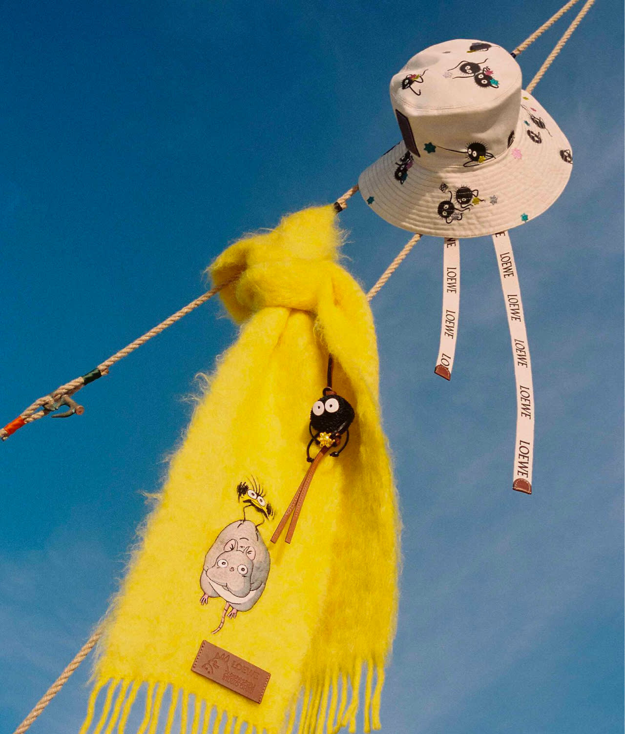A white bucket hat with cartoon characters and logo ribbons hangs on a rope next to a bright yellow fuzzy scarf adorned with cute character keychains, set against a clear blue sky.
