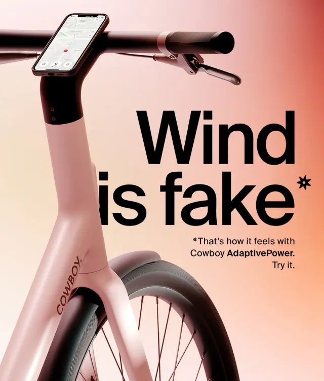 A sleek electric bicycle with a phone mounted on the handlebars is shown against a gradient background. Large text reads, Wind is fake* with smaller text below explaining Cowboy AdaptivePower technology.