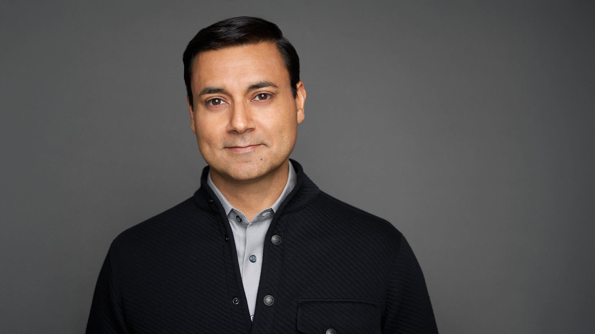 Baiju Shah - A man with short dark hair, wearing a light gray collared shirt and a textured black jacket, stands against a plain gray background, looking calmly at the camera.