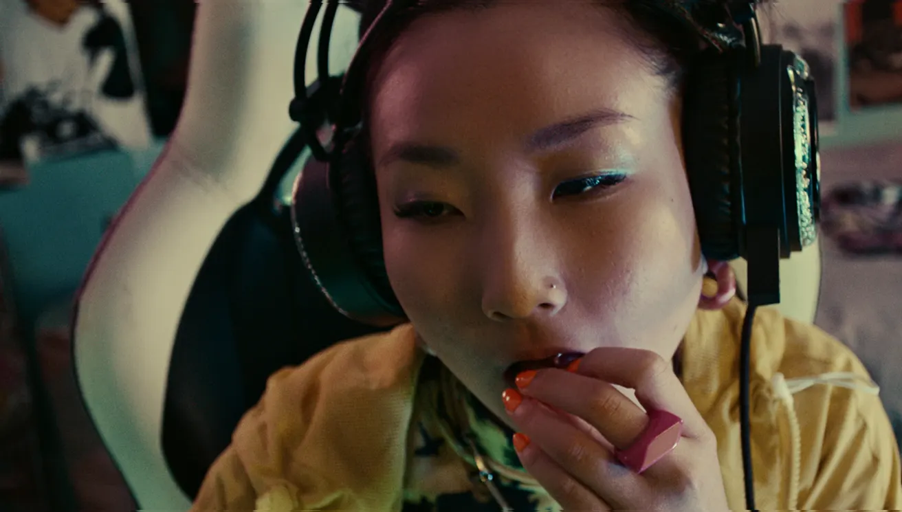A person with large headphones and bright makeup sits in a gaming chair, applying lipstick. Their nails are painted orange, and they wear a yellow top. The background is slightly blurred, suggesting a cozy room.