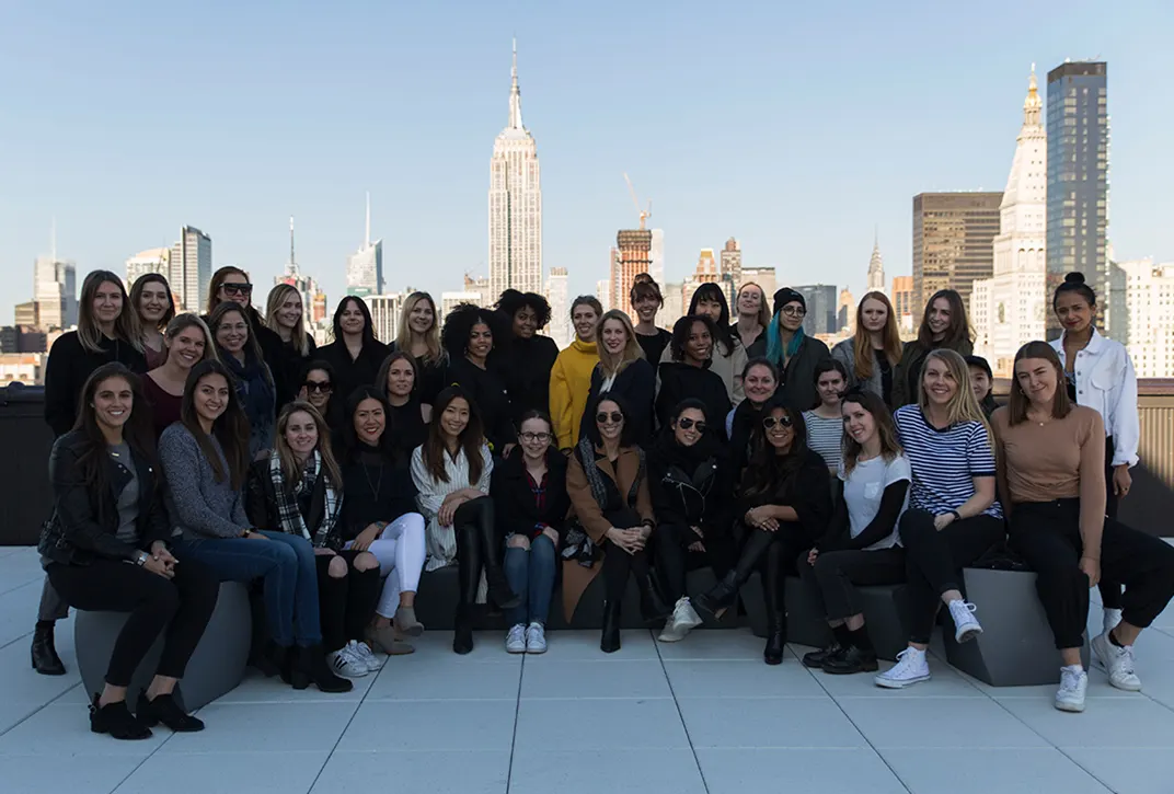 A large group of women poses together on a rooftop with a city skyline, including the Empire State Building, in the background on a clear day.