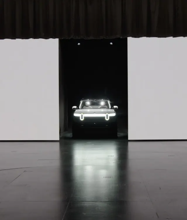 A vehicle with bright vertical LED headlights is emerging from a dark doorway in a large, mostly empty room with a reflective floor and a partially lowered curtain above.
