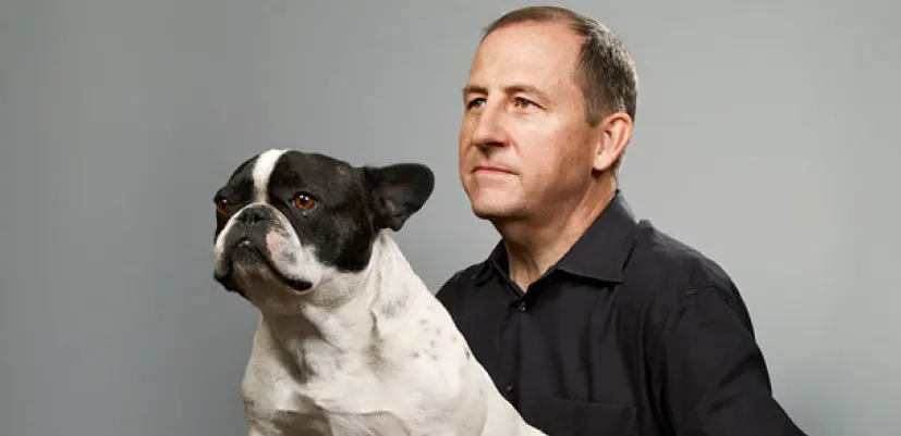 A man in a black shirt sits beside a black and white French bulldog, both looking intently in the same direction against a plain gray background.