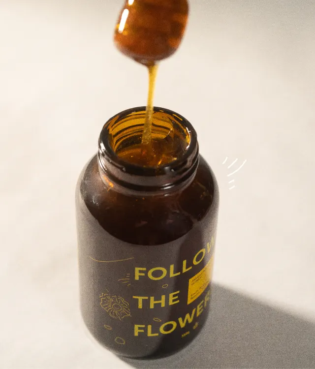 A spoon drizzles honey into an open brown jar labeled FOLLOW THE FLOWER against a light background.