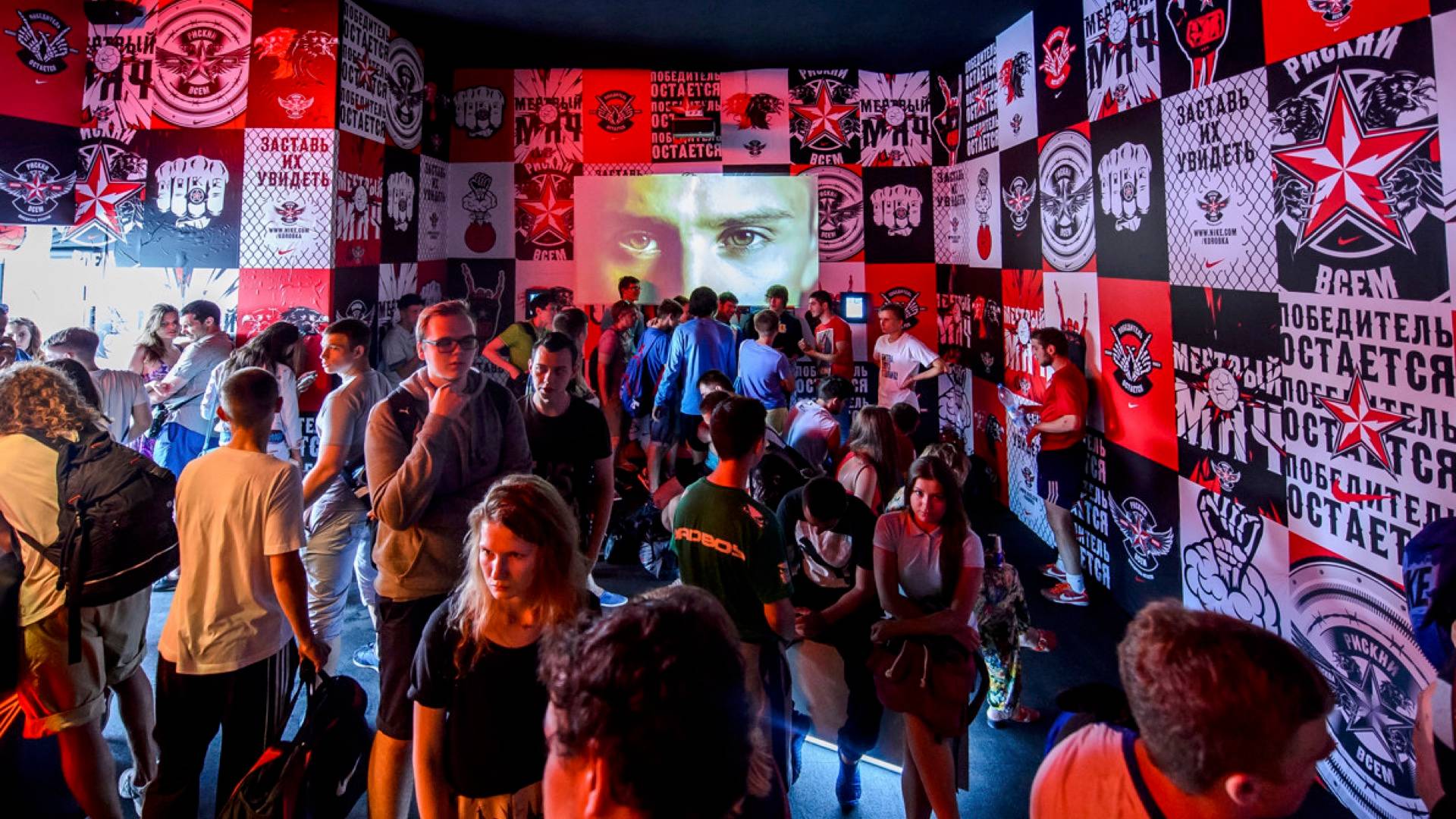 A crowd of people stands in a colorful, graffiti-covered room with large images and Russian text on the walls; a projected face with intense eyes is displayed above them, creating a vibrant and energetic atmosphere.