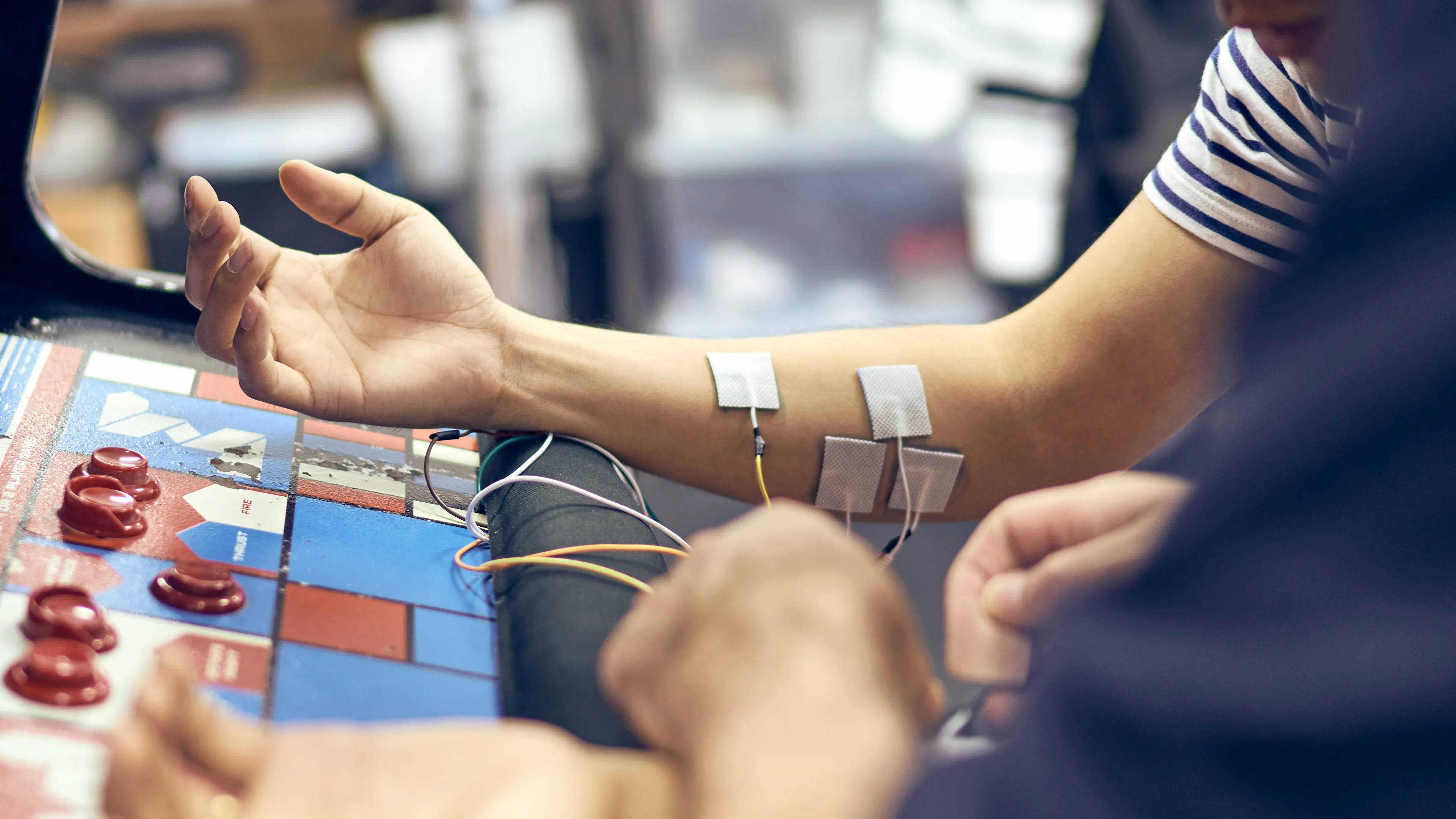 A person with electrodes attached to their forearm sits at a colorful table or arcade machine, while another person adjusts the wires connected to the electrodes.
