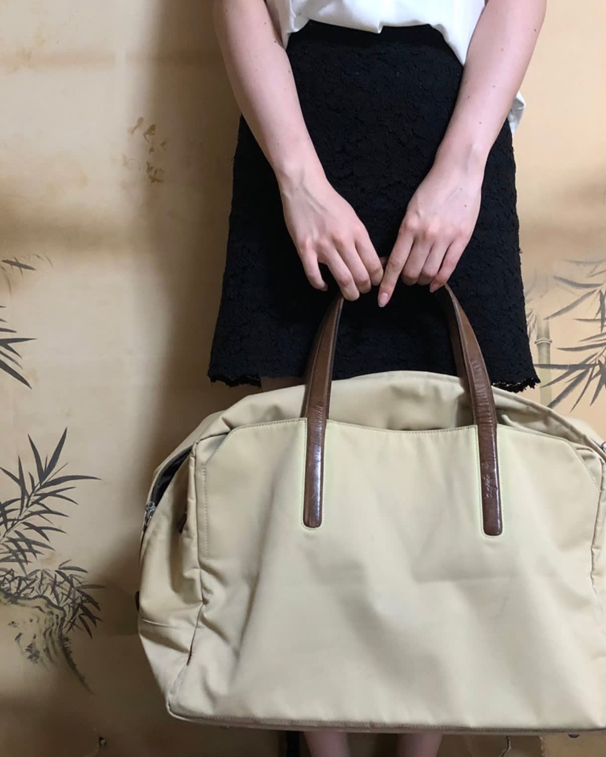 A person in a black skirt and white top is standing and holding a large beige bag with brown handles in front of them. Only the torso and arms are visible; the background features a subtle plant design.