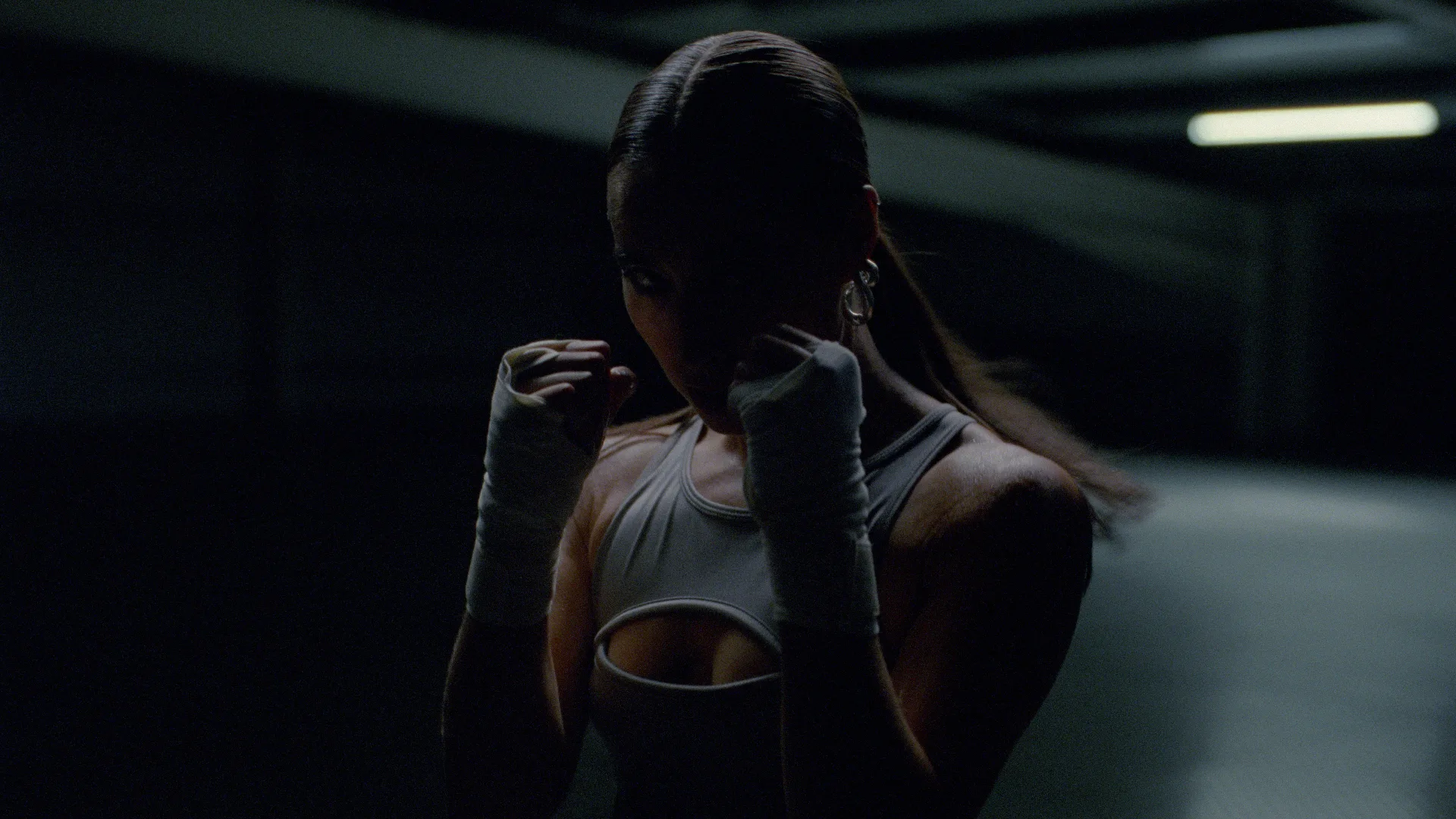 A woman with long hair in a ponytail stands in a fighting stance with her fists raised, wearing hand wraps and a sleeveless top, in a dimly lit, shadowy indoor space.