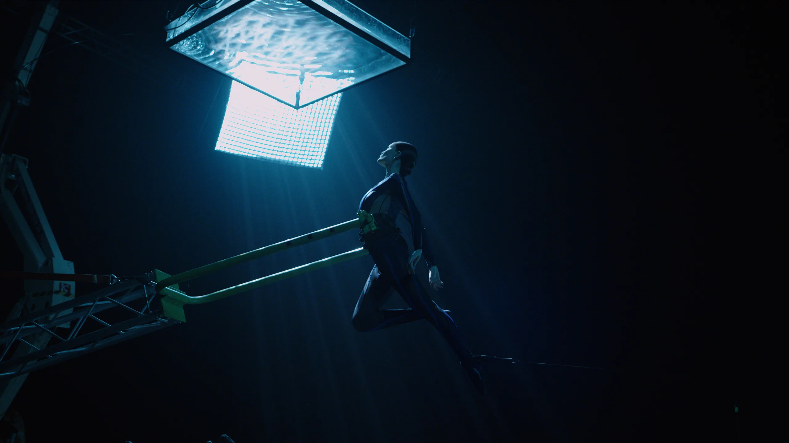 A performer in a dark bodysuit is suspended in midair by a harness, reaching toward a glowing, water-filled tank above, illuminated by dramatic blue lighting against a dark background.
