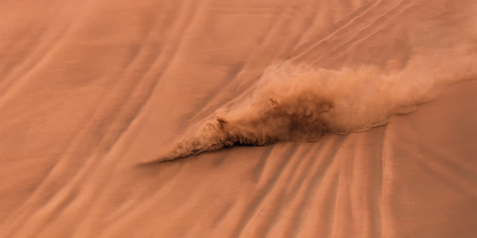 A plume of sand and dust rises as an object or animal quickly moves across smooth, orange sand dunes, leaving trailing lines and patterns behind.