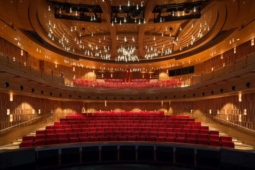 A modern, empty theater auditorium with multiple tiers of red seats, warm wooden walls, and an elaborate ceiling featuring dozens of hanging lights.
