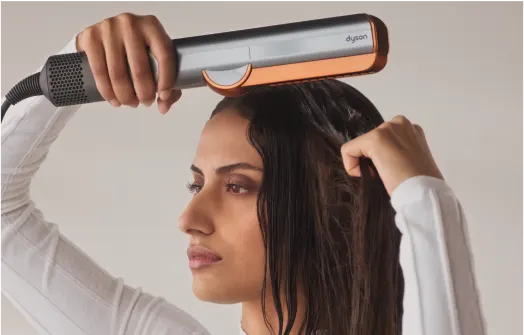 A woman with wet hair uses a Dyson hair straightener, gliding it over the top of her head. She wears a white long-sleeve top and looks focused on styling her hair.