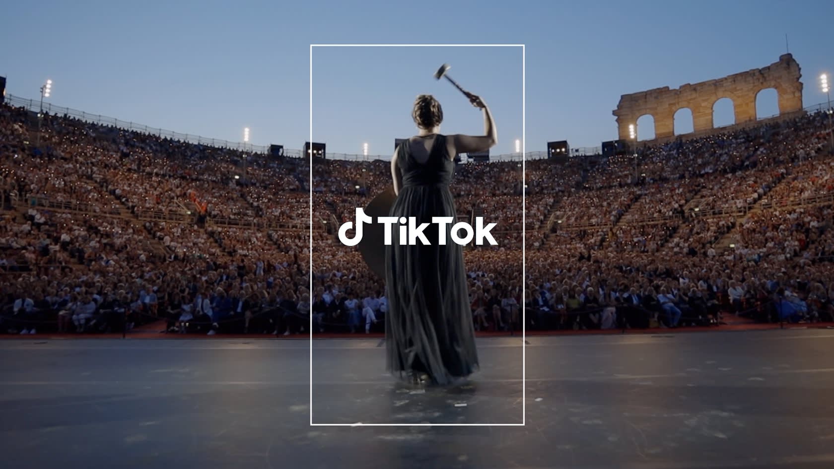 A woman in a black dress stands on stage facing a large outdoor audience, raising a gavel. The TikTok logo is overlaid at the center. An ancient coliseum is visible in the background at dusk.