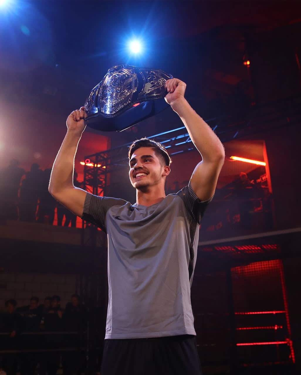A young man in a gray shirt smiles and holds a large silver championship belt above his head in a dimly lit arena with red lights. Spectators are visible in the background.