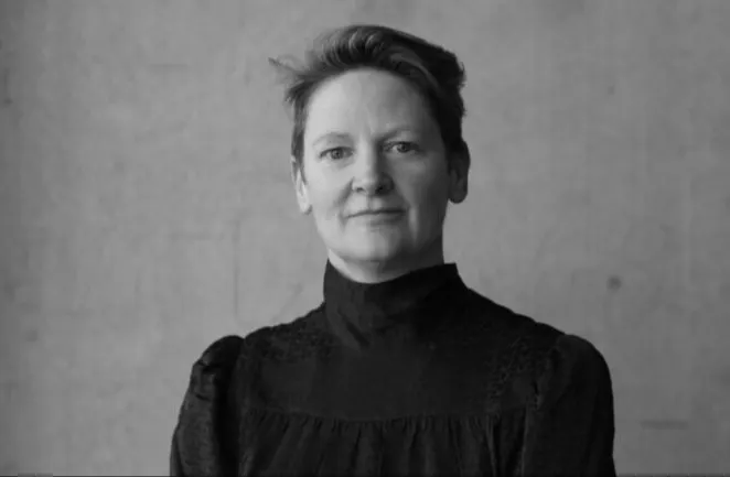 A person with short hair wearing a high-neck, long-sleeved dark top stands in front of a plain, light-colored background and looks directly at the camera, with a neutral expression. The photo is in black and white.