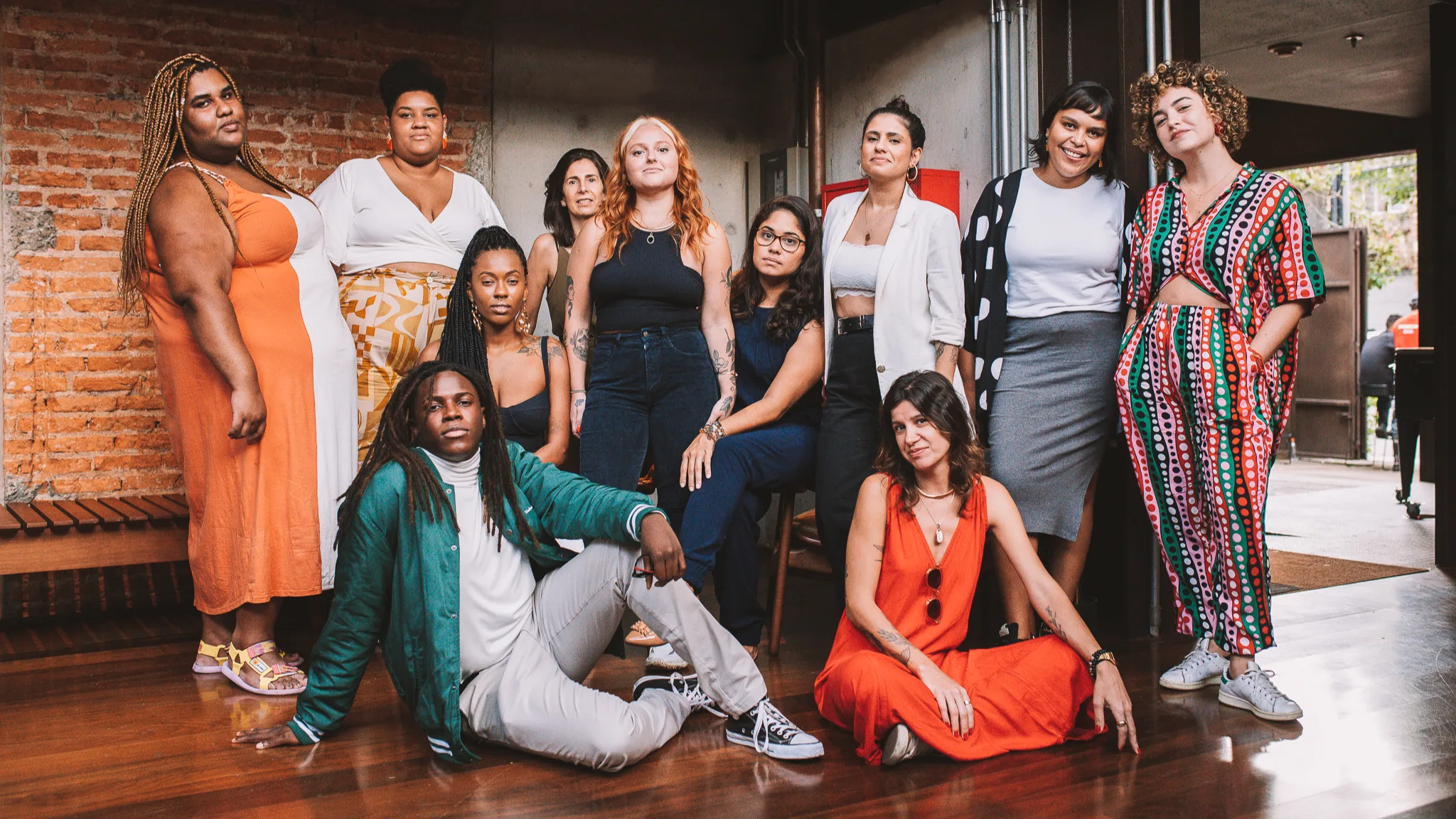 A diverse group of eleven people pose together indoors; some standing, others sitting or kneeling, all dressed in a variety of colorful and stylish outfits with a brick wall in the background.