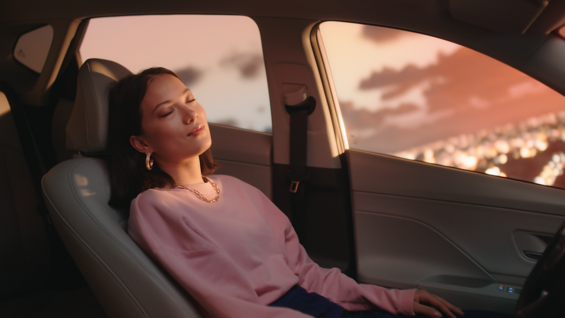 A woman reclines with her eyes closed inside a car, through the window a pink coloured sunset is visible with clouds and lights below.