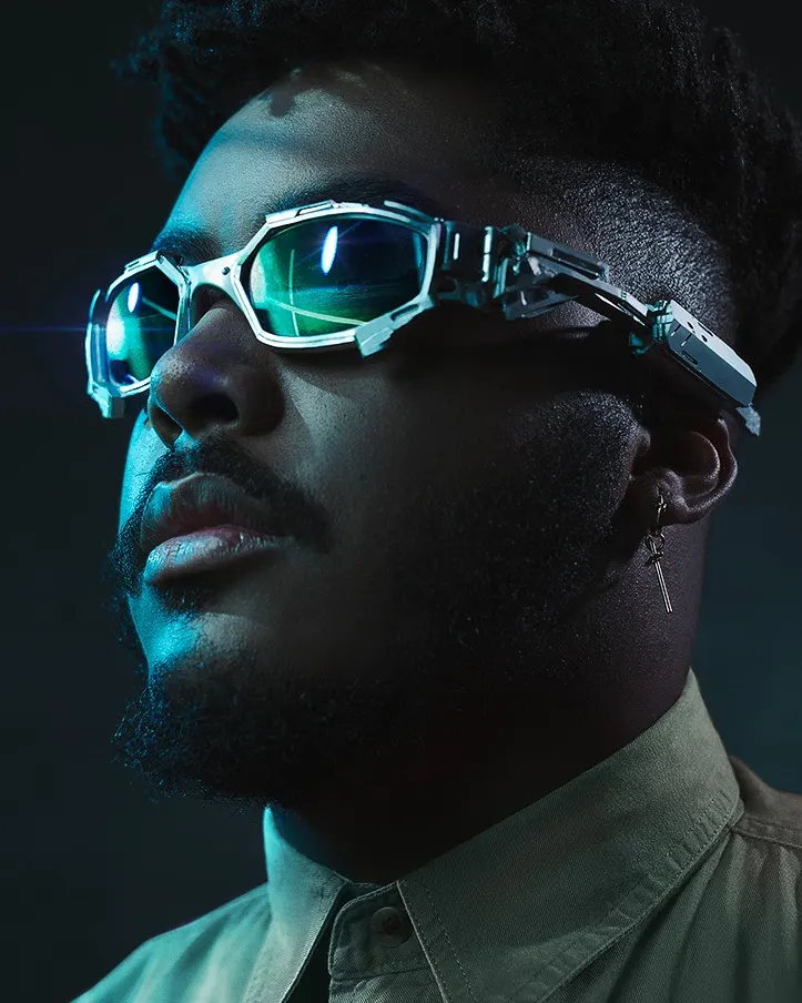 A man with a beard and mustache wears futuristic, reflective sunglasses and a green collared shirt. He has a cross earring in one ear and is lit with dramatic blue and green lighting against a dark background.