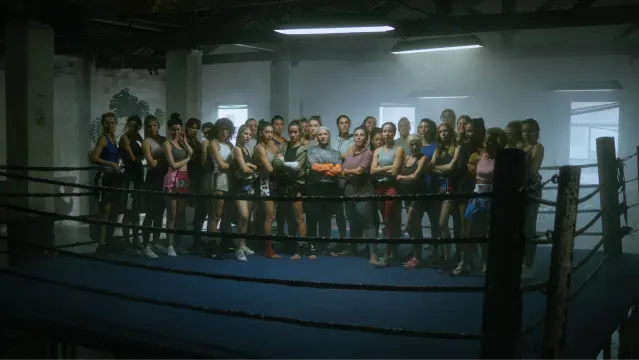 A large group of women in athletic wear stand together inside a boxing ring in a dimly lit gym, looking confidently toward the camera.