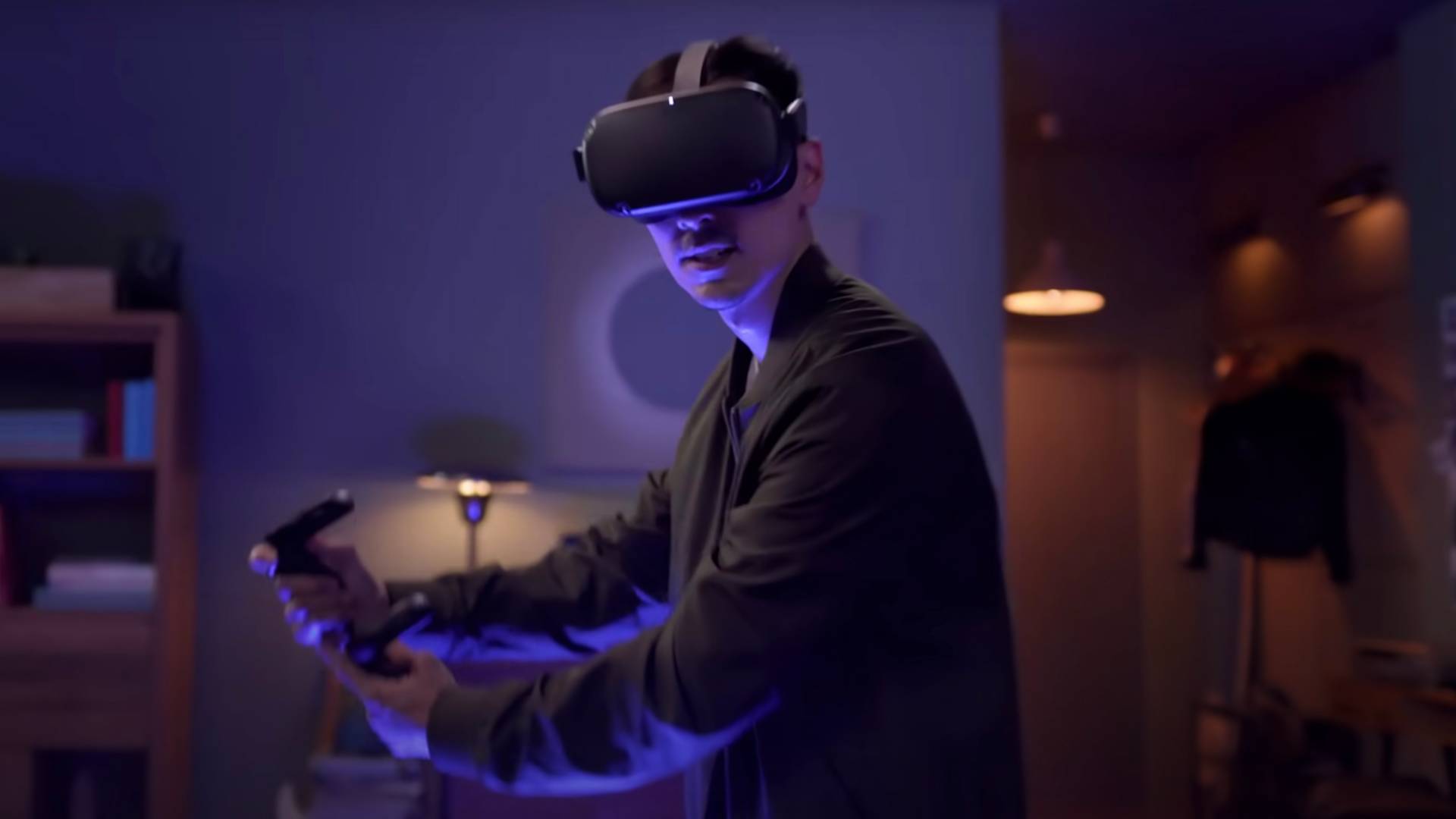 A person wearing a virtual reality headset and holding game controllers stands in a dimly lit room, illuminated by blue and purple lights, appearing focused and engaged in a VR experience.