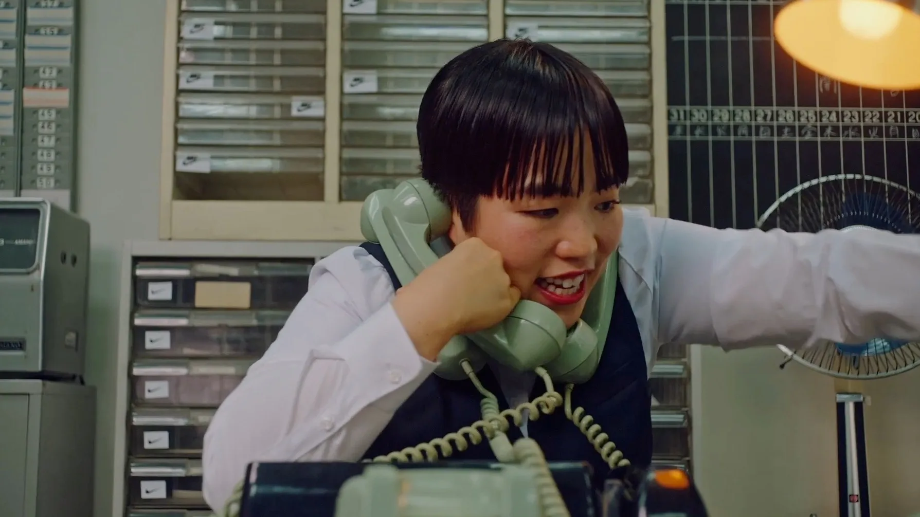 A person wearing a white shirt and dark vest is multitasking in an office, holding two corded phones to their ears while talking animatedly, surrounded by office equipment and organized shelves in the background.