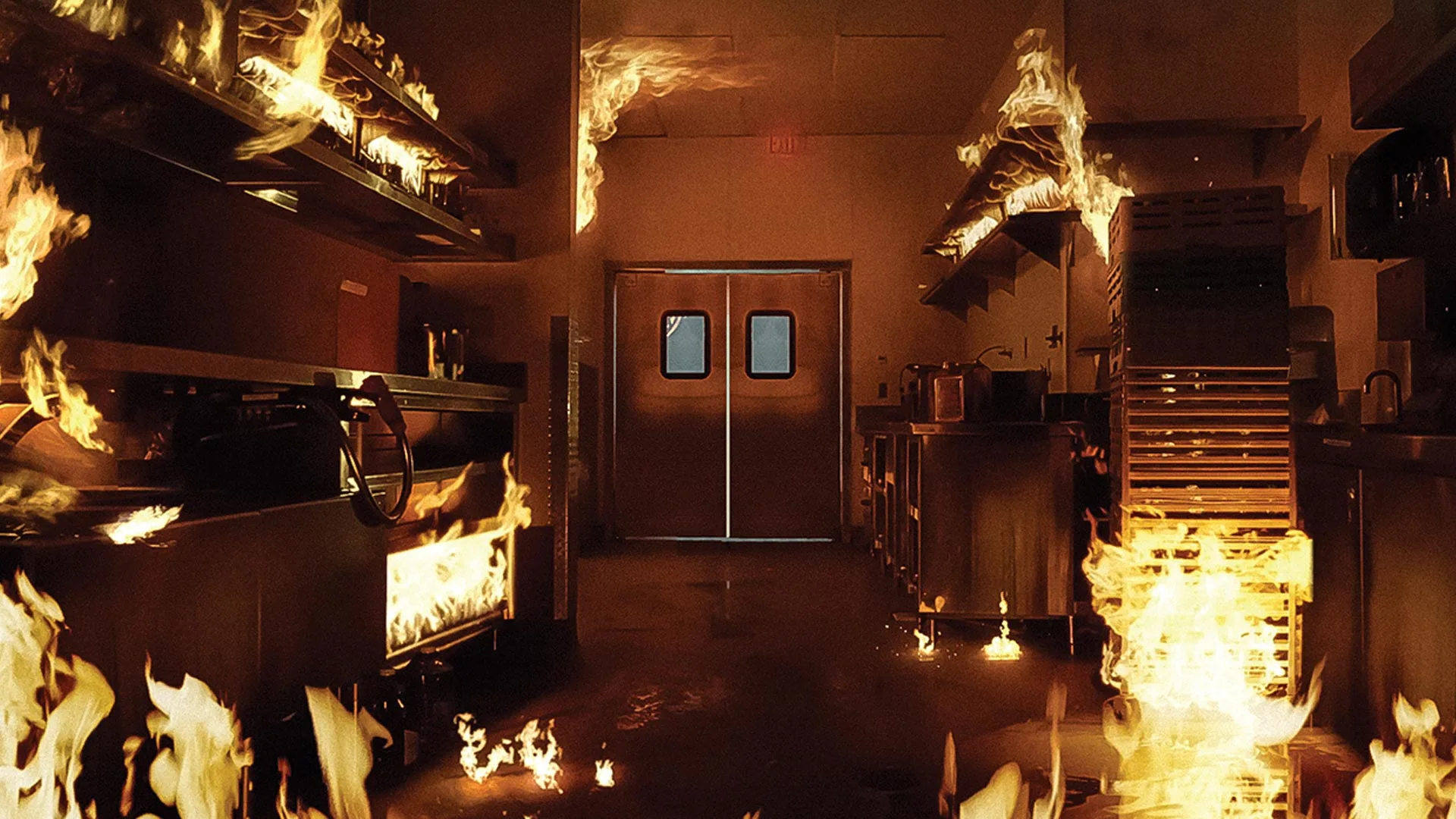 Flames spread throughout a commercial kitchen, with fire on counters, shelves, and the floor; double doors are visible at the back, and smoke fills the space, indicating a dangerous fire emergency.