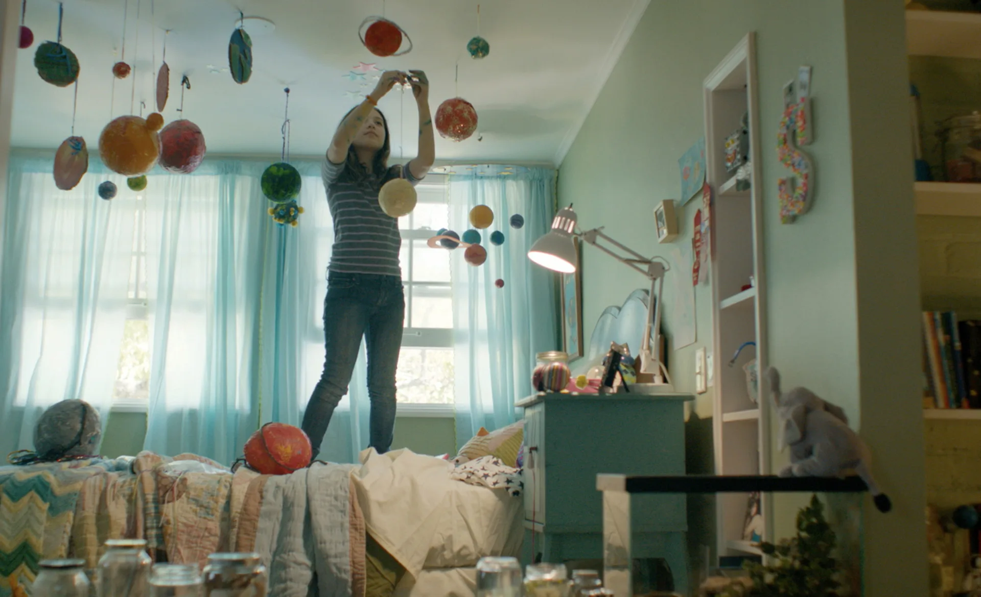A child stands on a bed, reaching up to adjust colorful hanging planets in a solar system mobile. The room is bright with turquoise curtains, books, and art supplies scattered around.