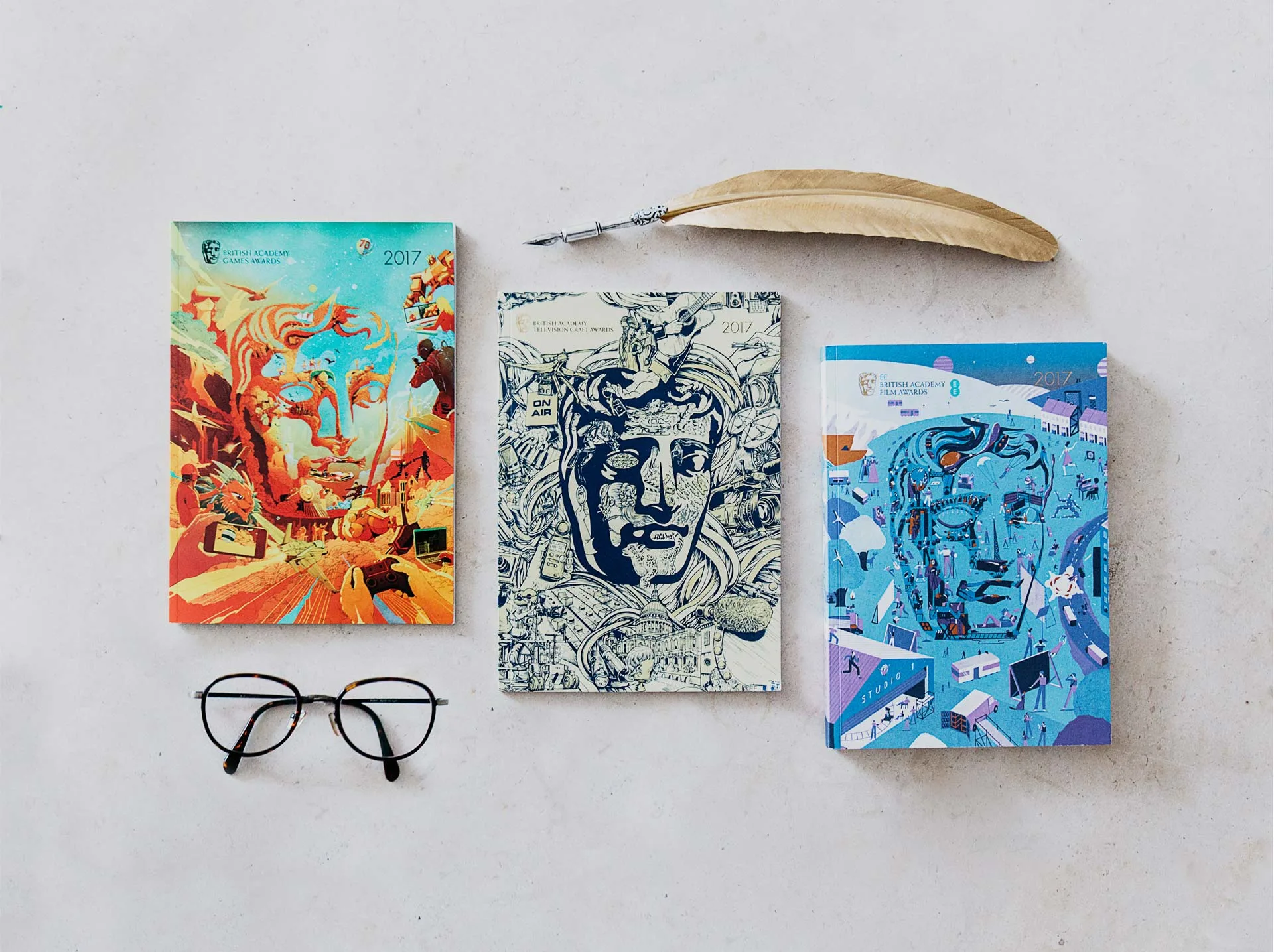 Three illustrated BAFTA notebooks with stylized faces are arranged on a light surface, accompanied by a quill pen and eyeglasses. The notebooks feature vibrant, artistic covers in orange, blue, and monochrome.