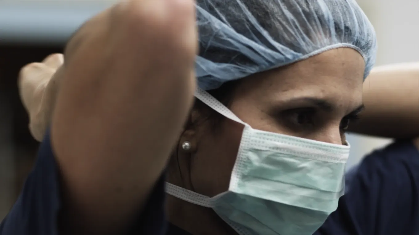 A person wearing a surgical mask and a blue hair cap adjusts their mask. The individual appears focused, preparing for a medical procedure or work in a healthcare setting.