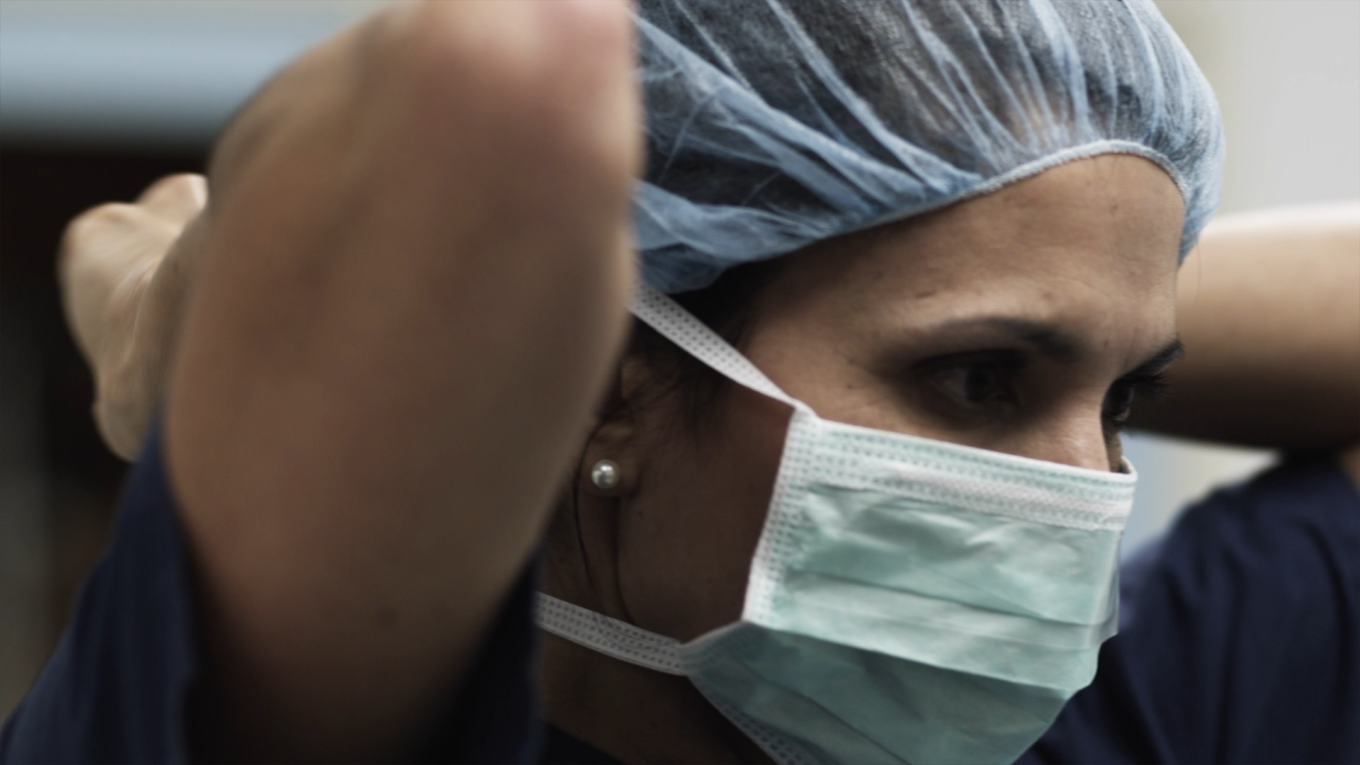 A person wearing a surgical mask and a blue hair cap adjusts their mask. The individual appears focused, preparing for a medical procedure or work in a healthcare setting.