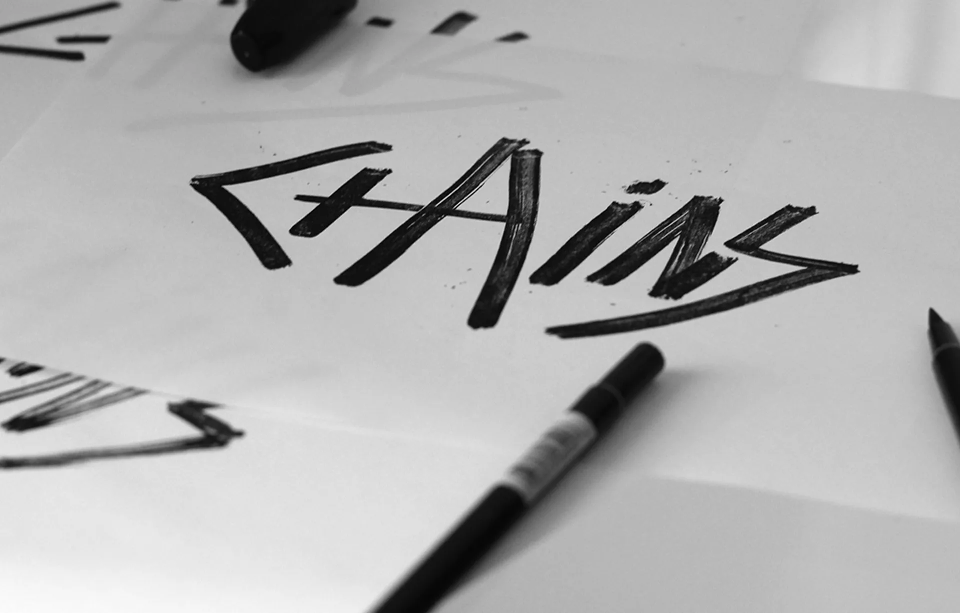 A piece of paper with the word CHAINS written in bold, black, jagged letters. Black markers are scattered around the paper, and part of another sheet with similar writing is visible.
