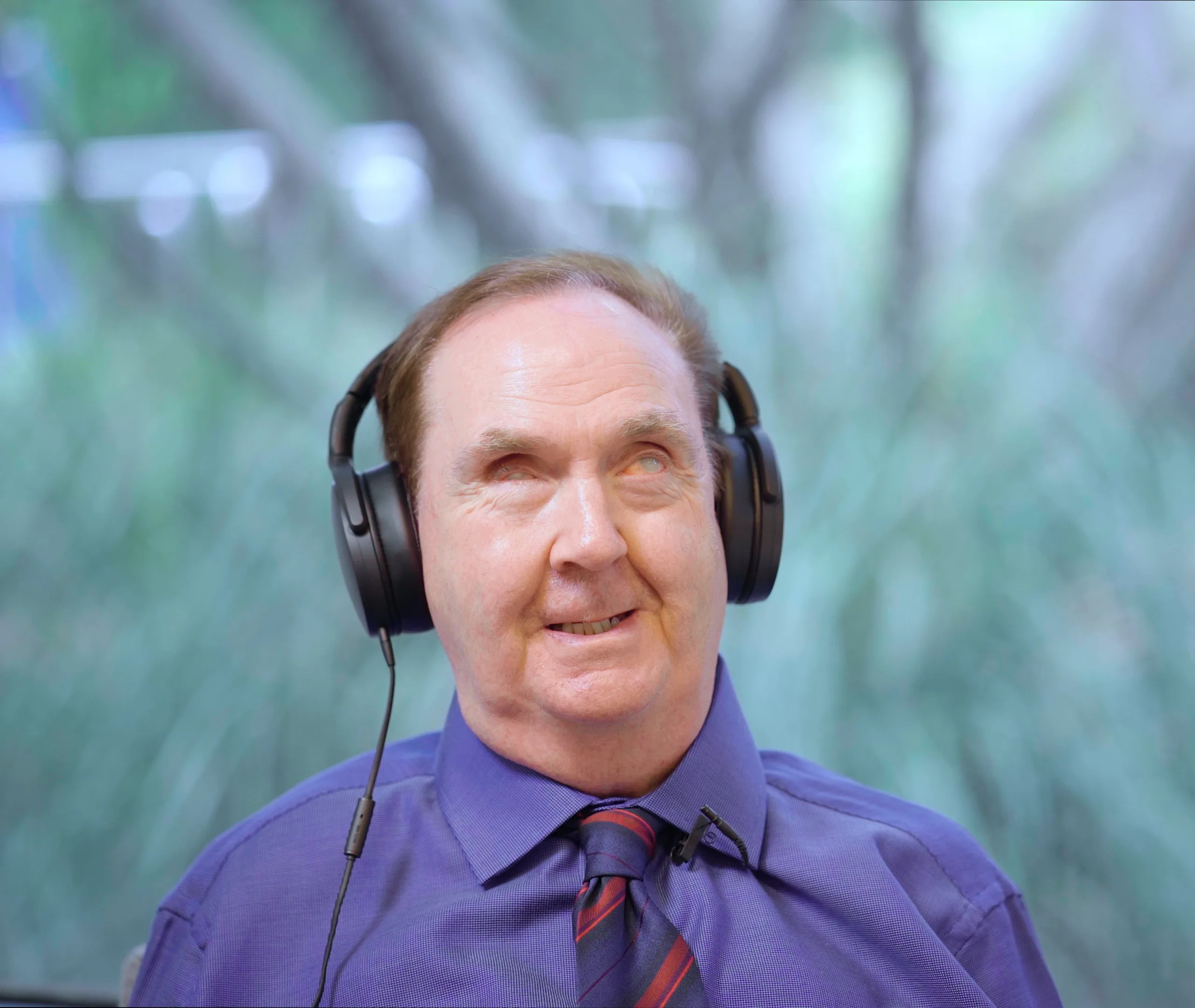 A man wearing a purple shirt, striped tie, and large black headphones is smiling while looking slightly upward. The blurred background suggests an outdoor, natural setting with greenery.