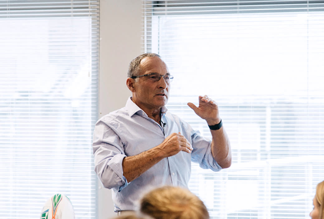 A man wearing glasses and a light blue shirt gestures while speaking to a group in a bright room with large windows and closed blinds.