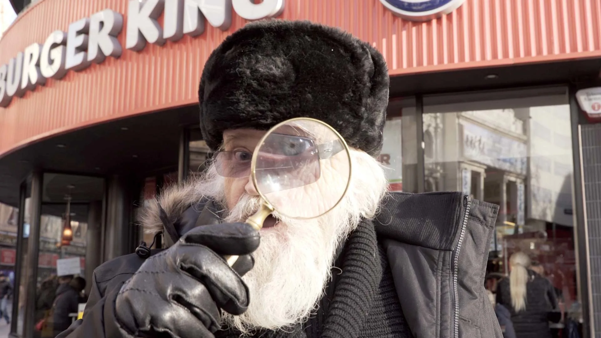 An older man with a white beard and black fur hat holds a magnifying glass up to his face, standing in front of a Burger King restaurant. He wears black gloves and a dark coat.