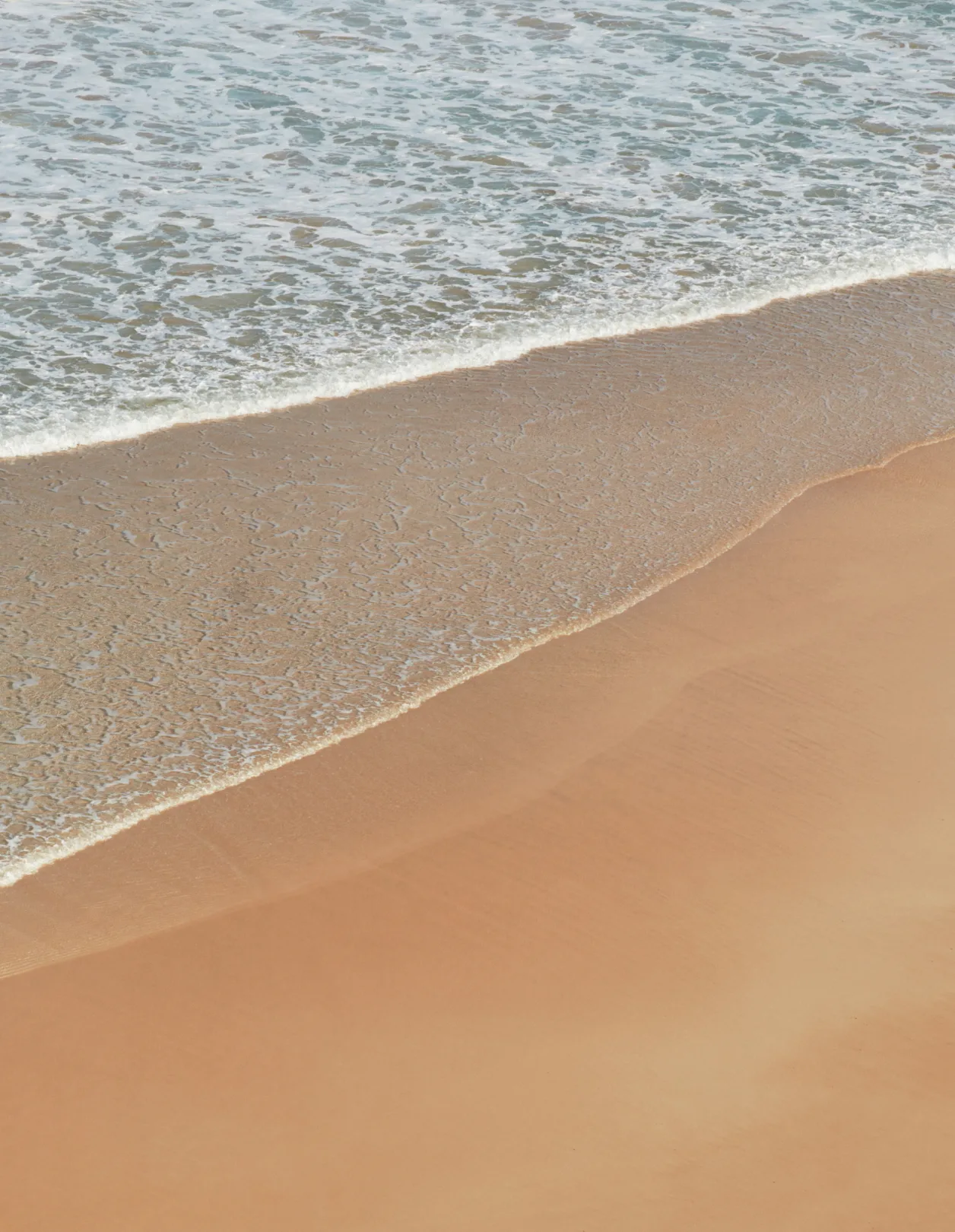 A gentle wave washes onto a smooth sandy beach, creating soft patterns where the water meets the shore under natural light.