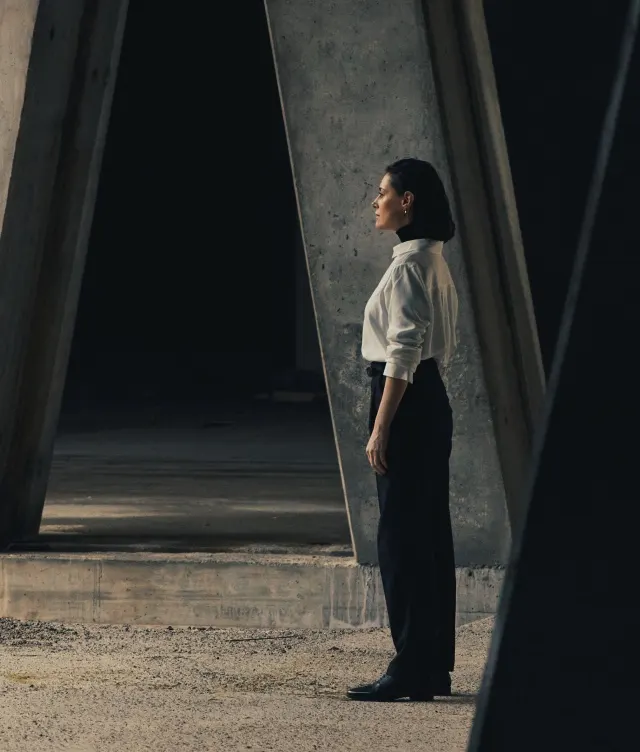A woman in a white shirt and black pants stands in profile under large, angular concrete columns in a dimly lit, industrial setting.