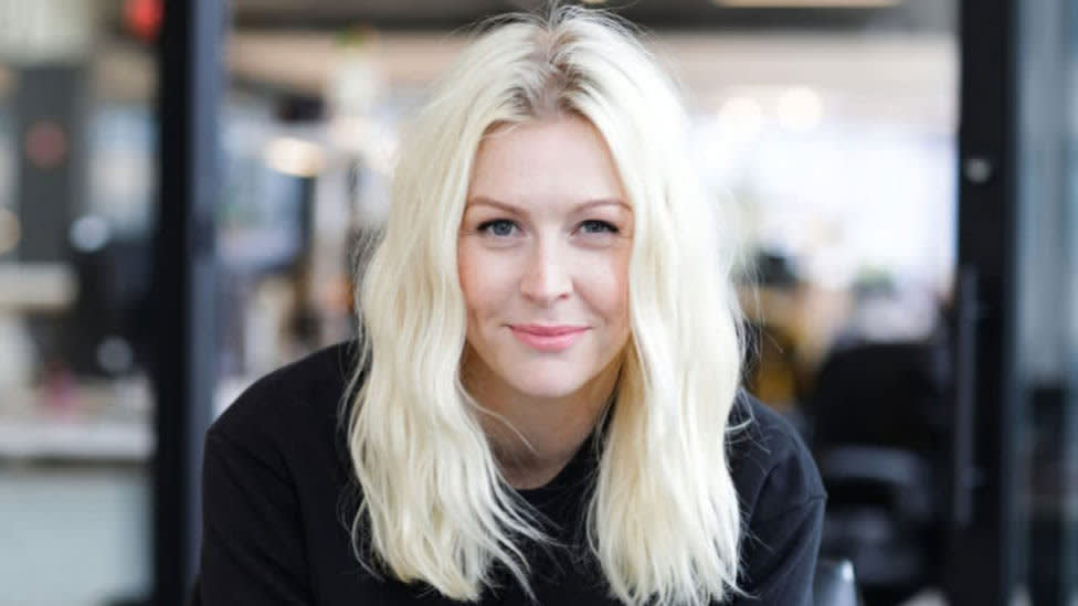 A woman with long, wavy blonde hair and a black top is smiling softly at the camera in an office setting with blurred background.