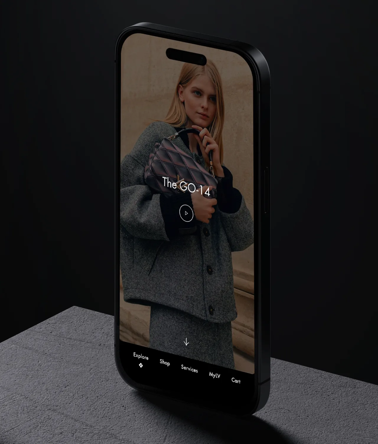 Phone displaying a woman in a casual grey jacket and matching skirt, she is holding a small blue and pink quilted handbag.